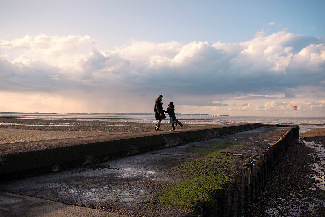 Happy couple in winter coats on sunny ocean beach jetty