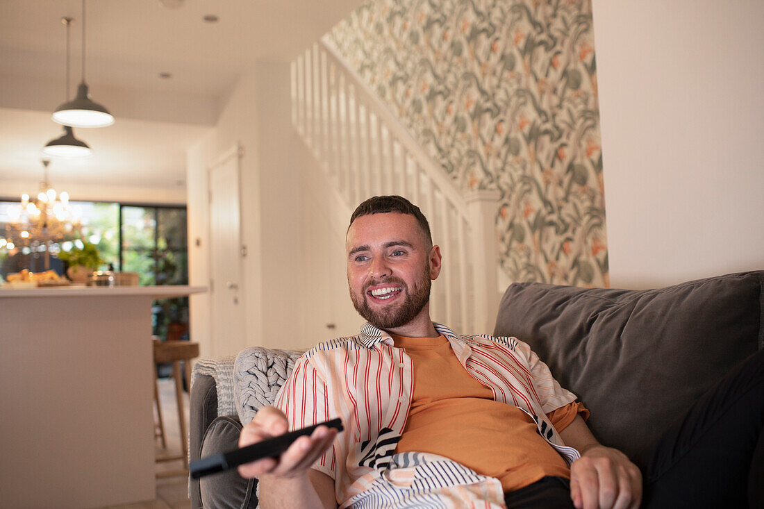 Happy young man with remote control watching TV on sofa