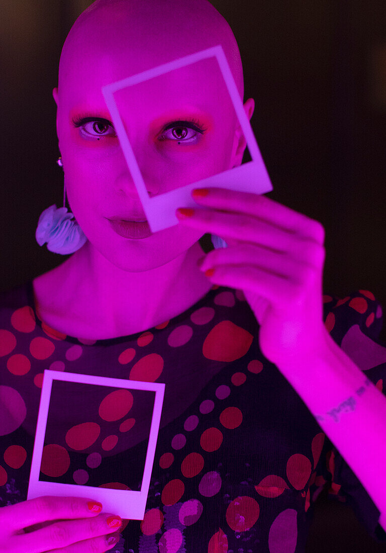 Woman with shaved head holding polaroid over eye