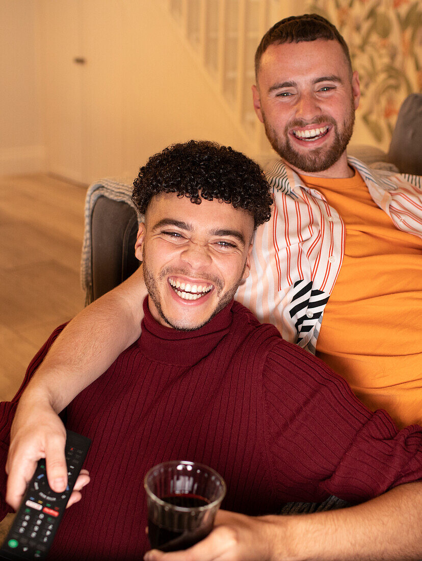 Happy gay couple watching TV and drinking red wine at home