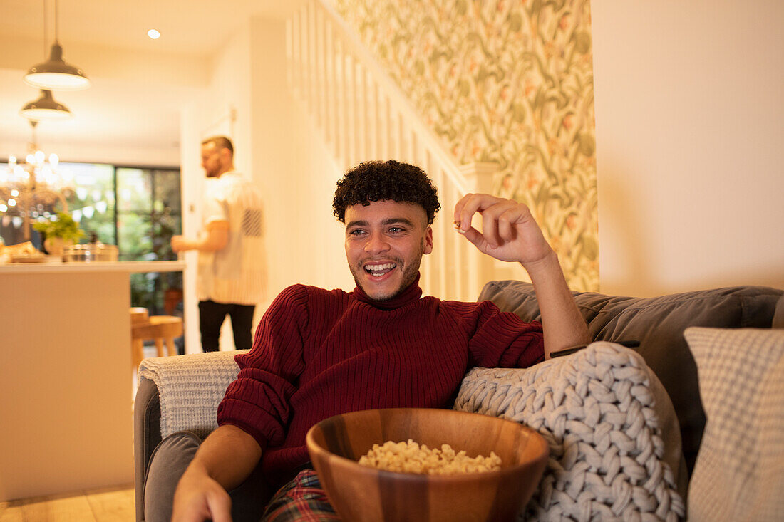 Happy young man with popcorn watching TV on living room sofa