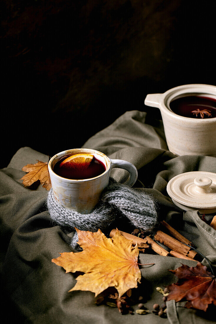 Hot mulled wine in ceramic pot and mug in scarf with spices, orange and autumn leaves over dark linen tablecloth