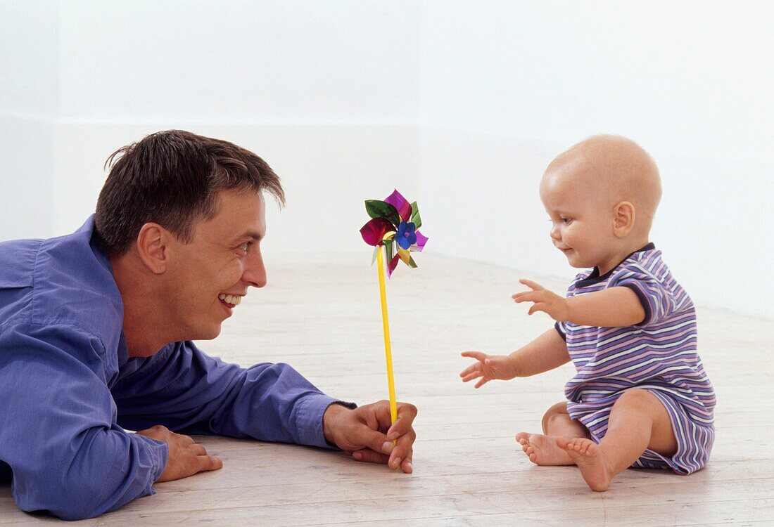 Man lying on the floor holding paper windmill towards a baby