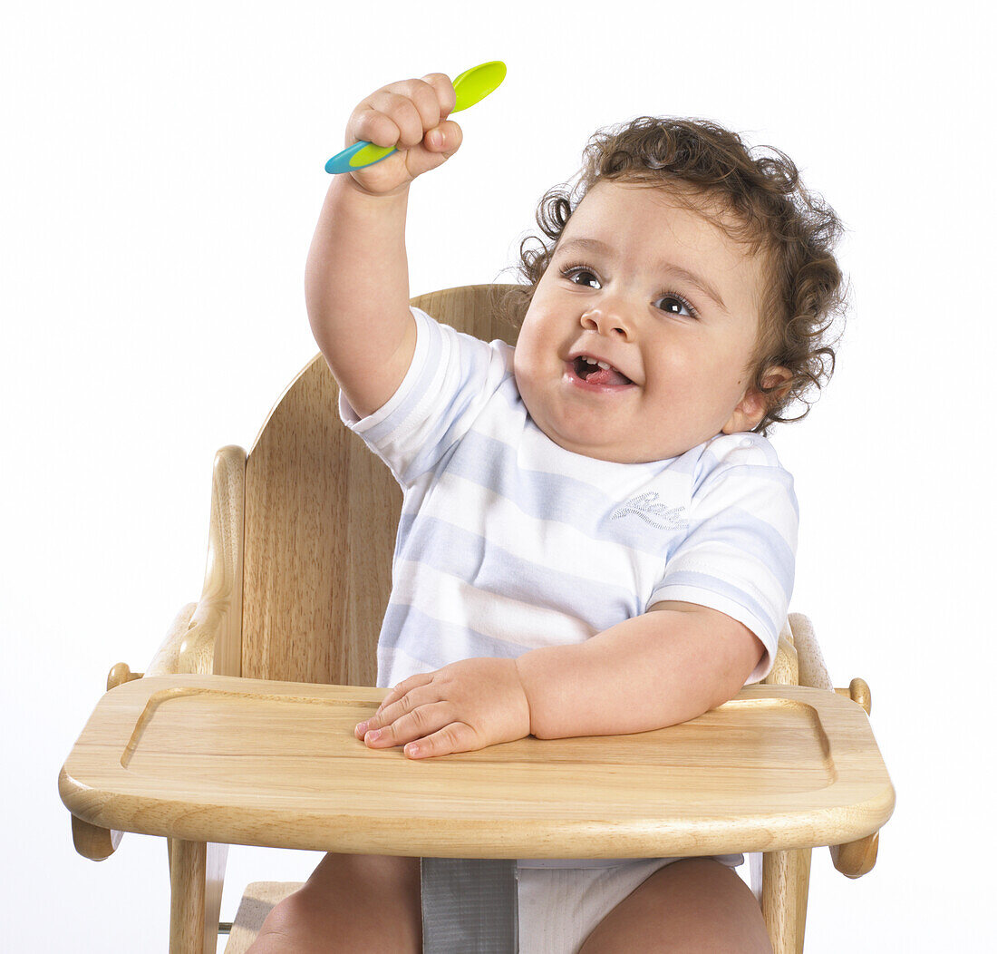 Baby boy sitting in high chair, holding up spoon