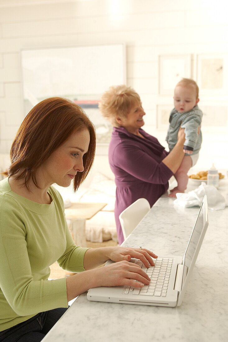 Mother using laptop as grandmother holds grandson