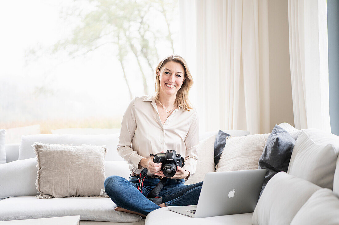 Blonde woman sitting on a couch with photo camera and laptop