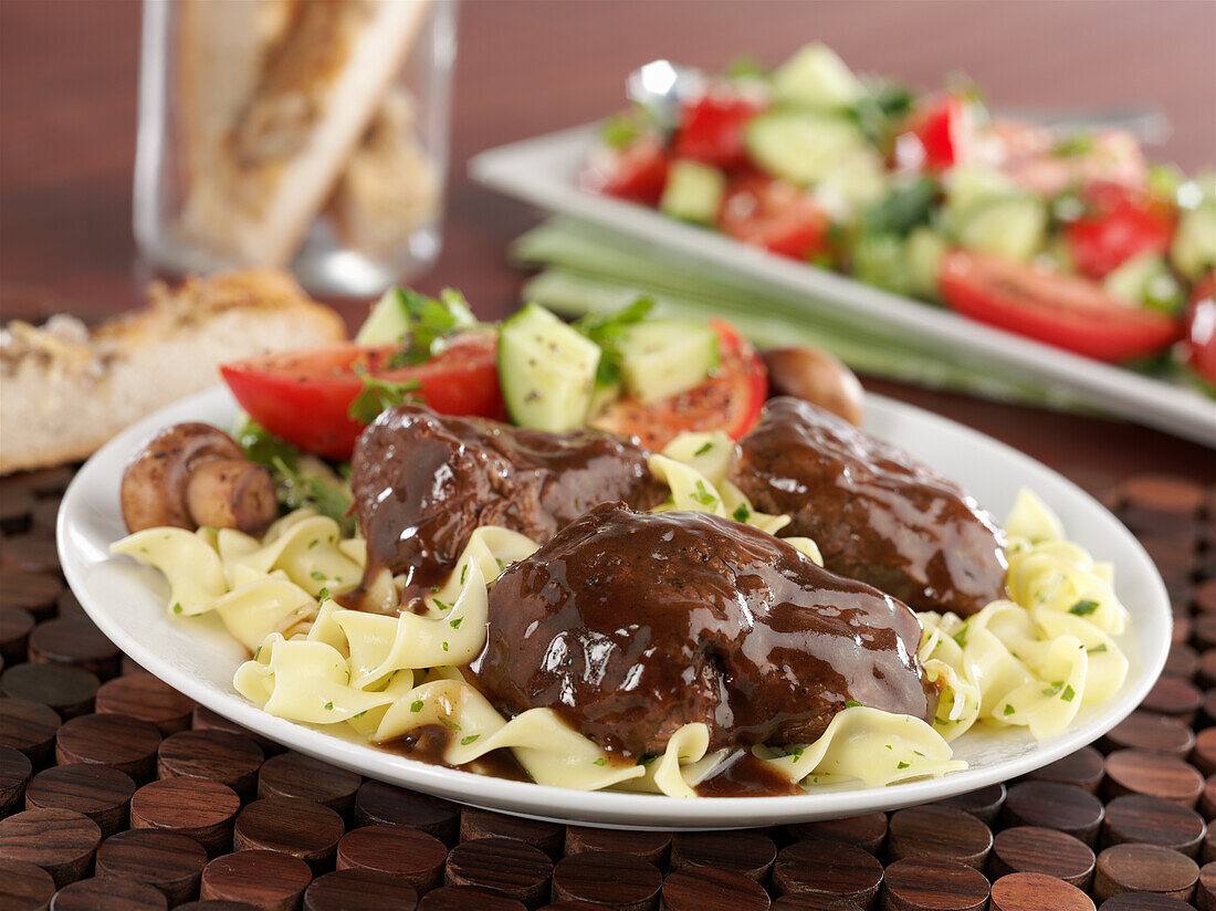 Braised beef short ribs with braising liquid on a bed of pasta with cucumber and tomato salad