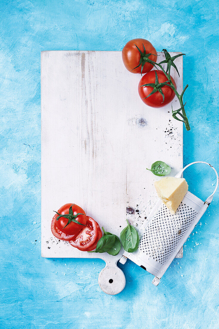 Weißes Holzbrett, Tomaten, Basilikum, Käsereibe auf blauem Untergrund