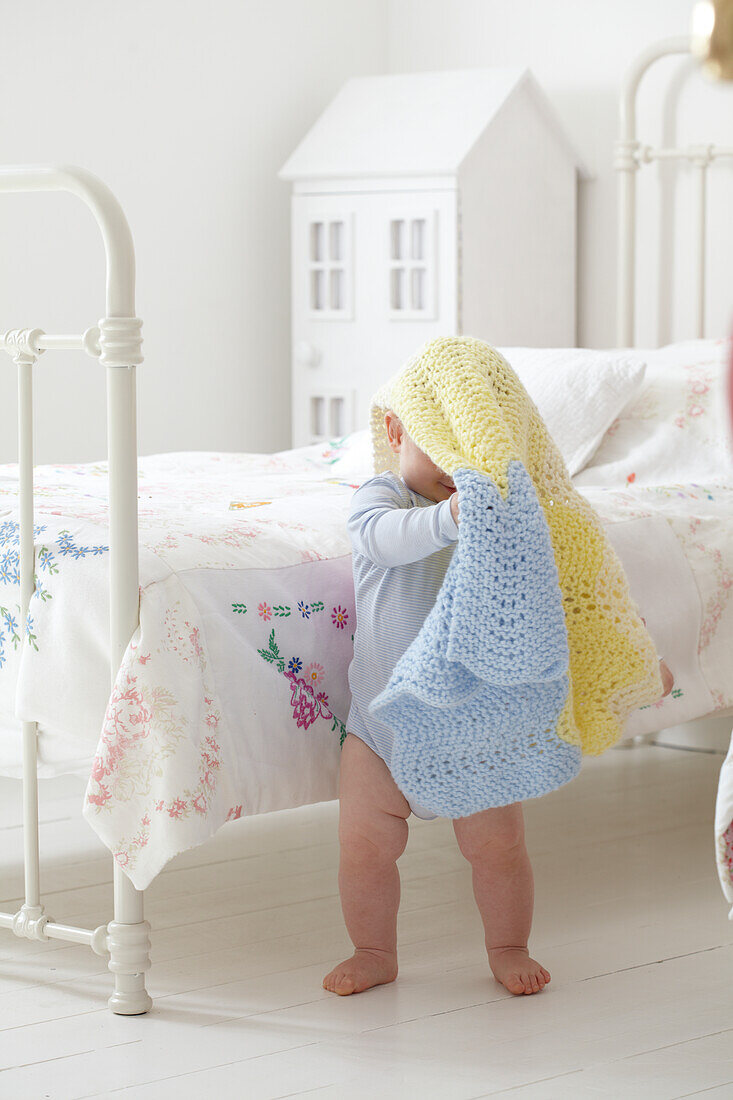 Baby boy standing next to bed with blanket over his head