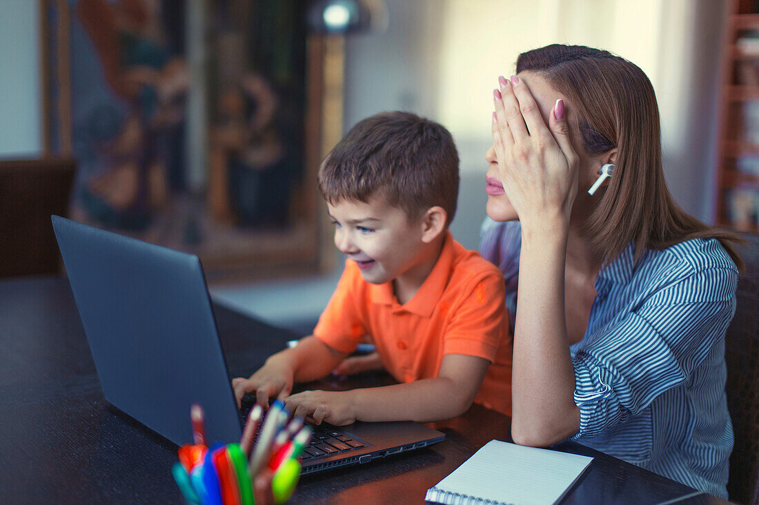 Tired woman working whilst son types on a laptop