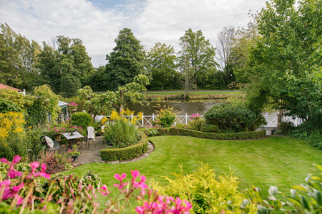 Gepflegter Garten mit Blumen, Buchshecken und Rasenfläche, dahinter Fluss