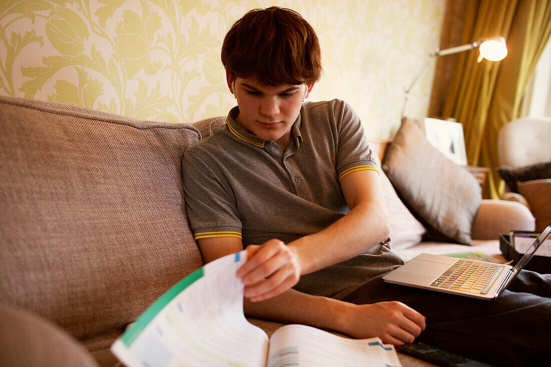 Teenage boy with laptop and textbook studying at home