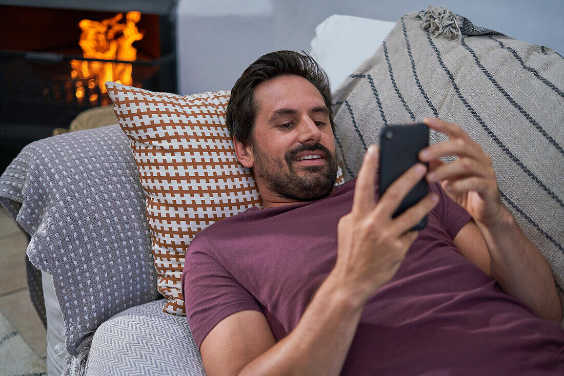 Man with smartphone relaxing on living room by fireplace