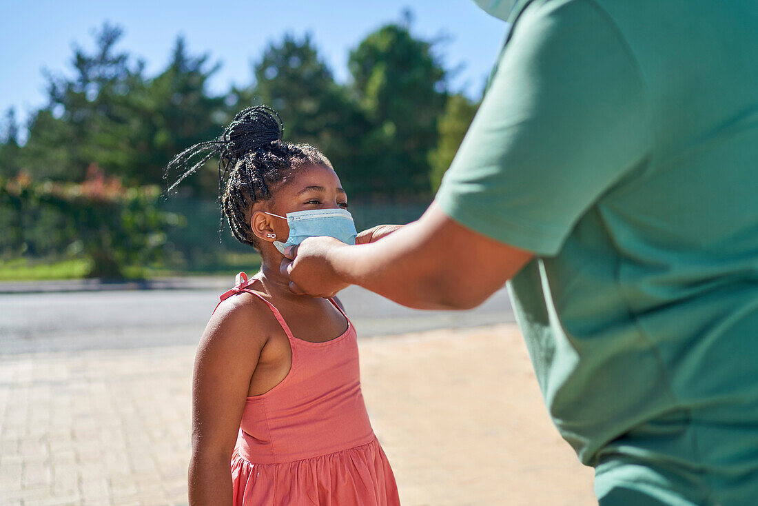 Father helping daughter with face mask on sunny sidewalk