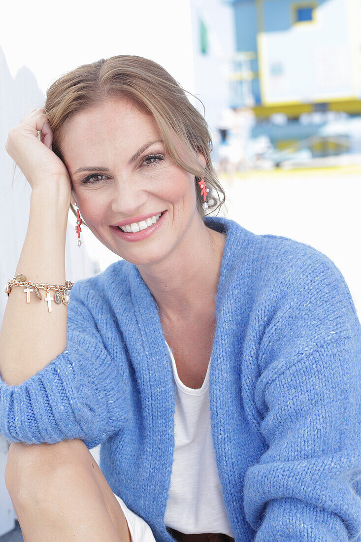 Langhaarige Frau in weißem Sommerkleid und blauer Strickjacke am Strand sitzend