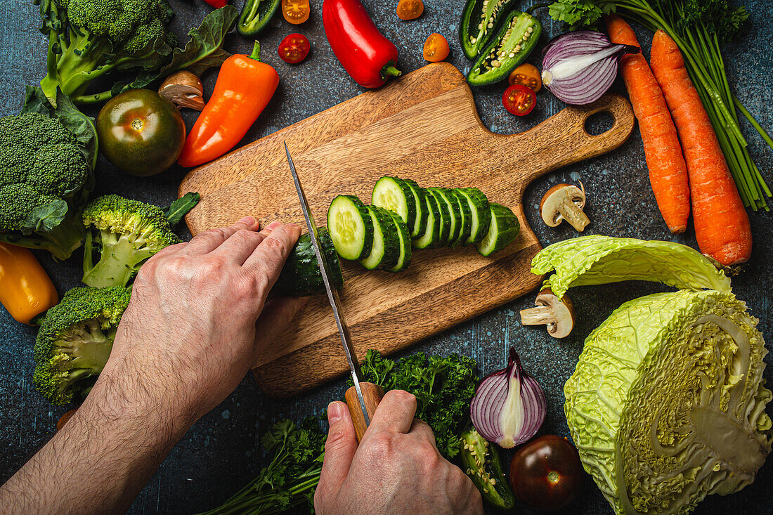 Hands chopping cucumber on wooden cutting board and assorted fresh vegetables