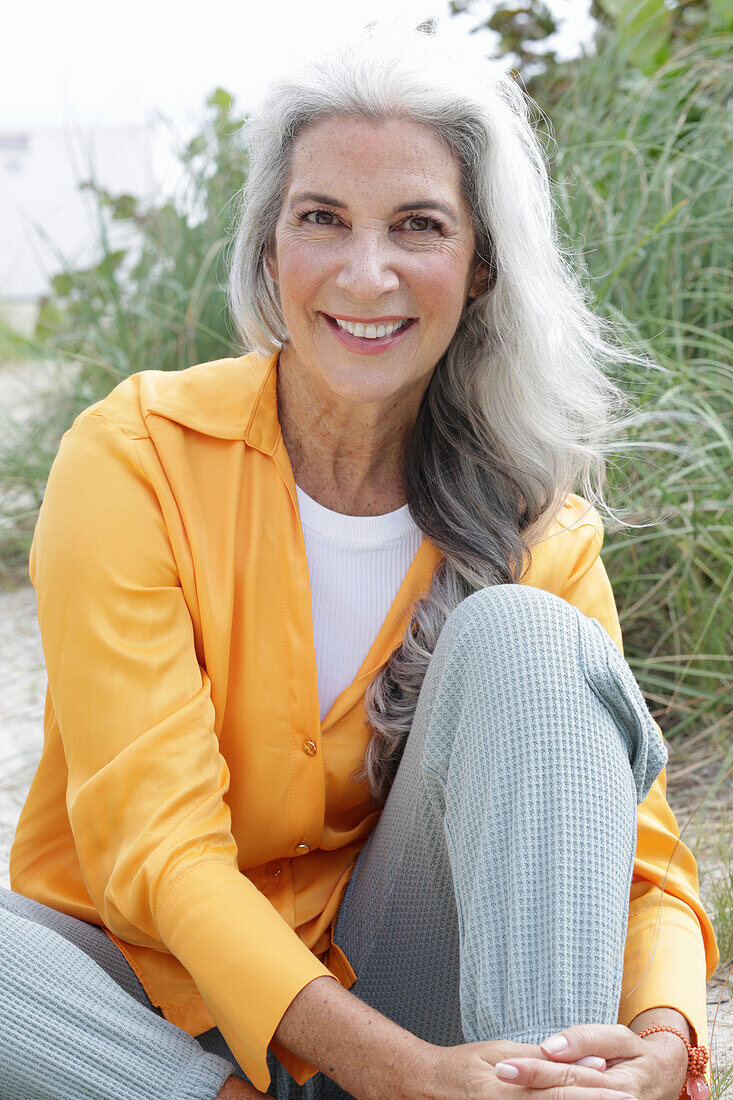 Mature woman with grey hair in orange blouse and trousers sitting on the beach