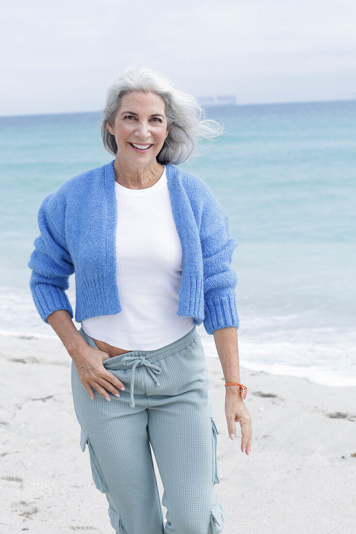 Mature woman with grey hair in white t-shirt, blue cardigan and trousers on the beach