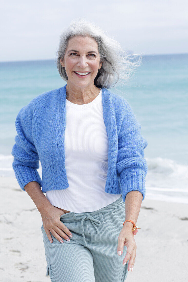 Reife Frau mit grauen Haaren in weißem T-Shirt, blauer Strickjacke und Hose am Strand