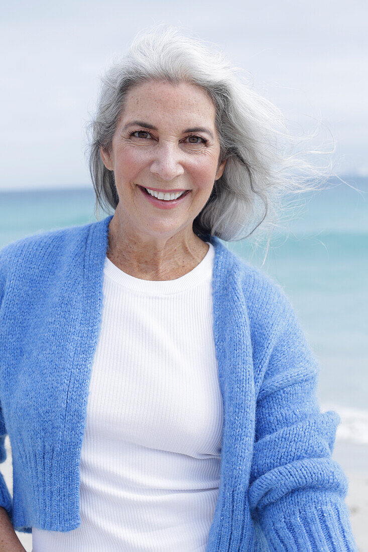 A mature woman with grey hair wearing a white t-shirt and a blue cardigan on a beach