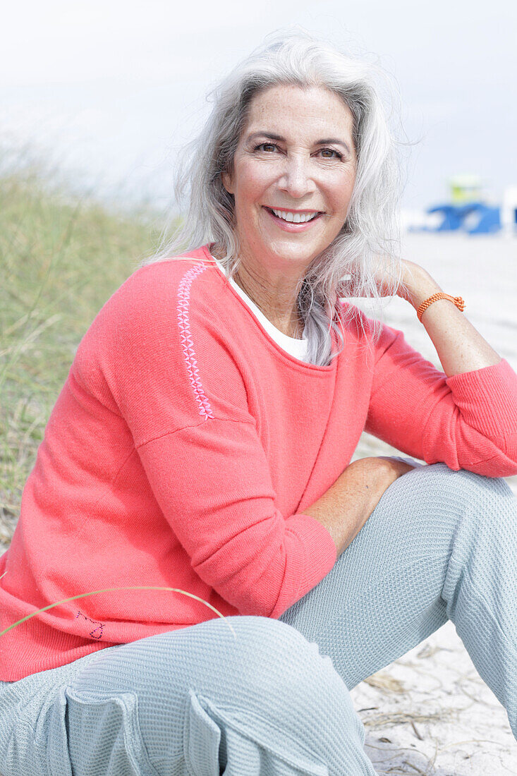Reife Frau mit grauen Haaren in lachsfarbenem Pullover und Hose am Strand