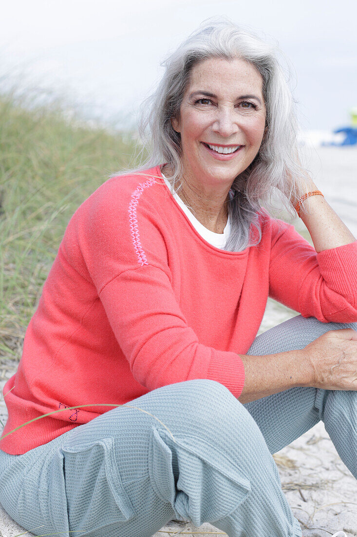 Reife Frau mit grauen Haaren in lachsfarbenem Pullover und Hose am Strand