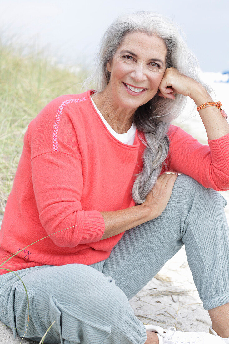 Reife Frau mit grauen Haaren in lachsfarbenem Pullover und Hose am Strand