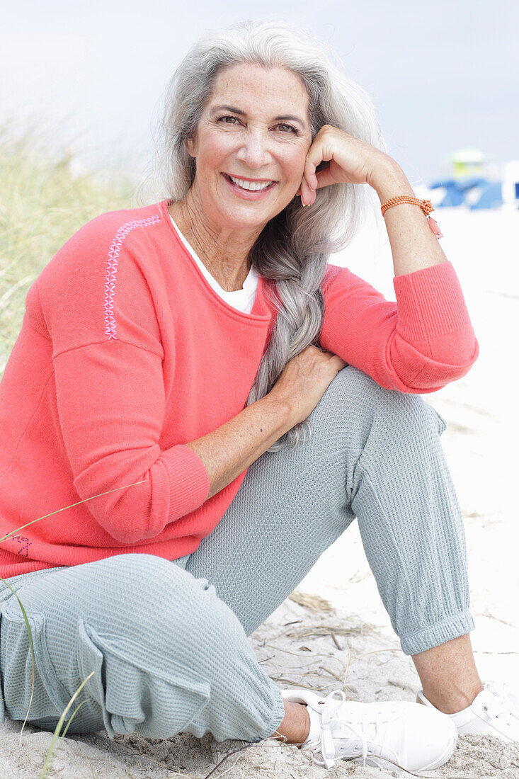 Reife Frau mit grauen Haaren in lachsfarbenem Pullover und Hose am Strand