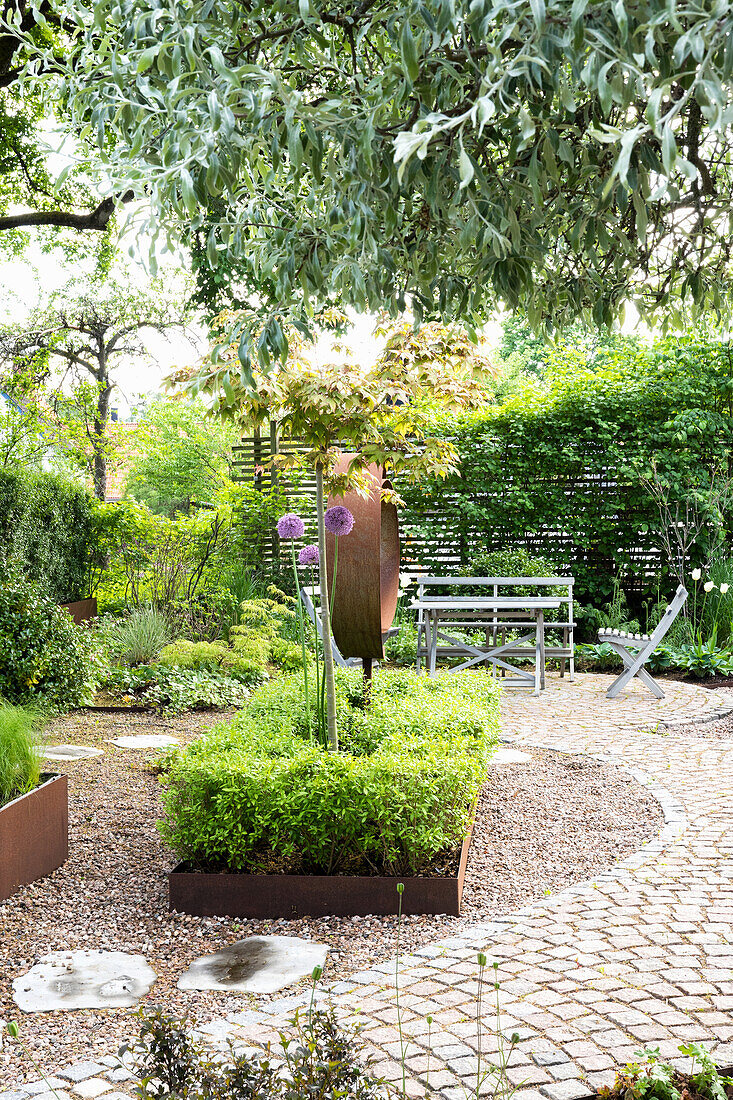 Gartenweg aus Pflastersteinen, eingefasstes Beet mit niedriger Hecke, Zierlauch und Skulptur aus Metall mit Edelrost