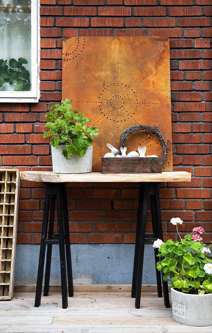 Ornaments and geraniums on and around old table on wooden terrace against brick wall