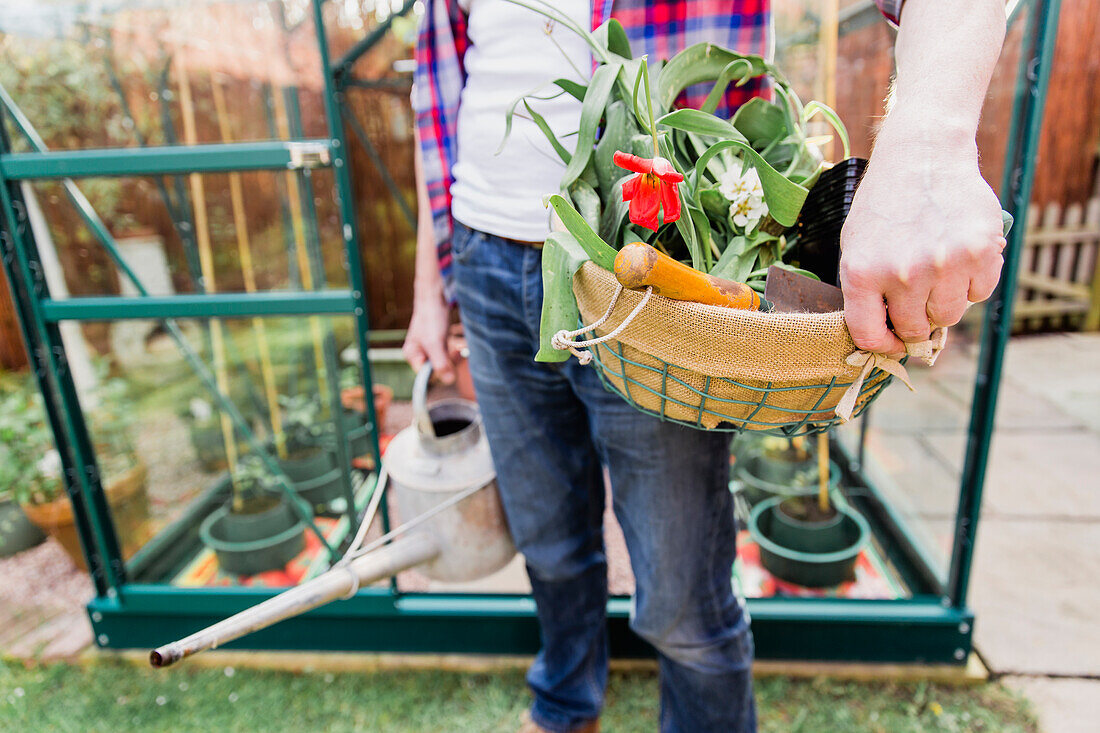 Close-up of man holding basket with flowers and watering can before greenhouse in garden