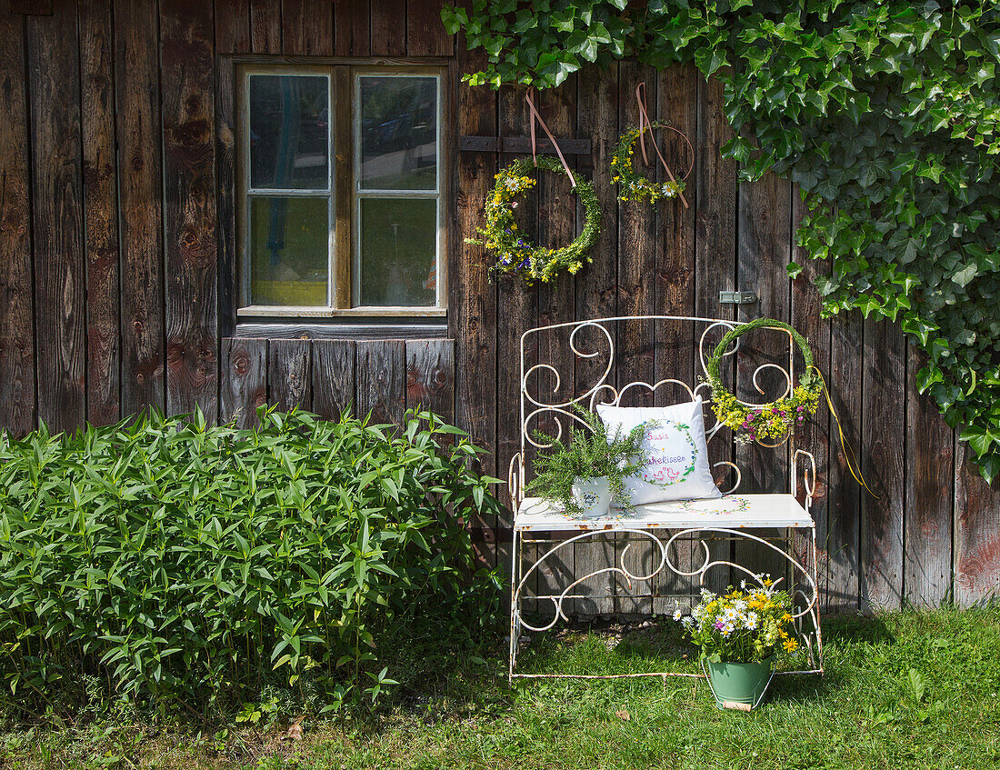 Gartenbank mit Kissen, Blumenstrauß und Blumenkränze an Holzwand