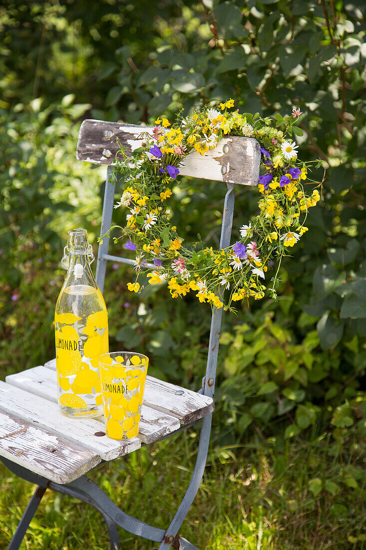 Blumenkranz auf altem Holzstuhl mit Limonadenflasche und Glas im Garten