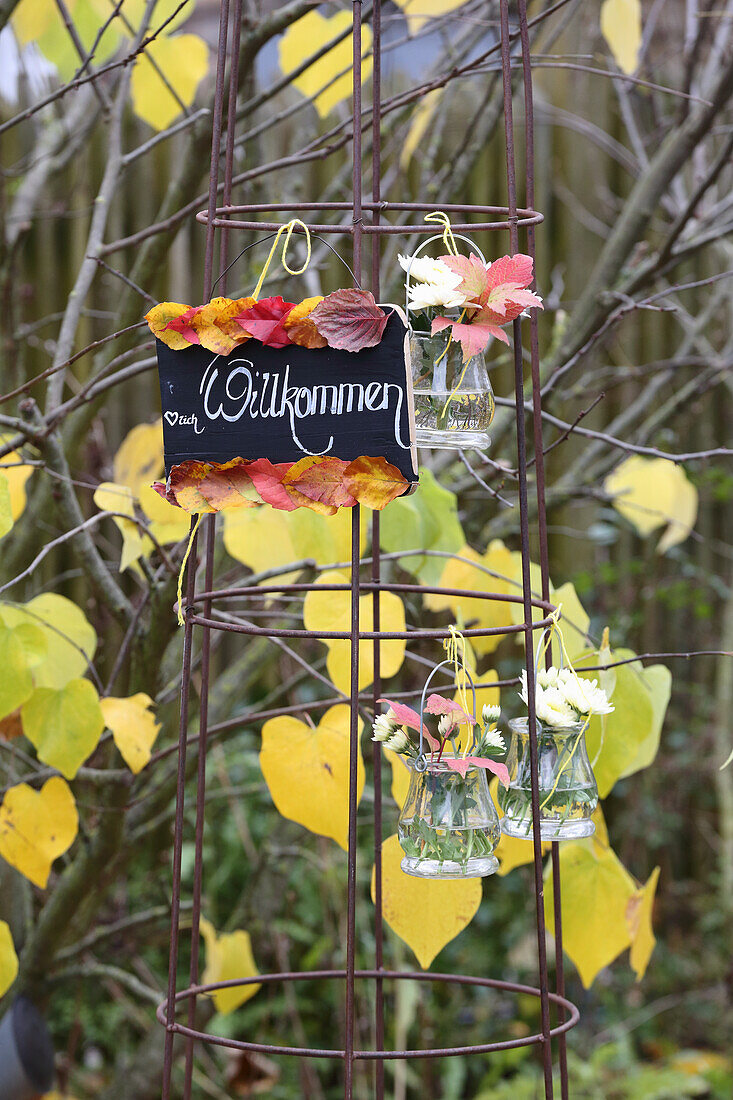 Willkommensschild, Herbstblätter und Hängevasen an Rankhilfe im Garten