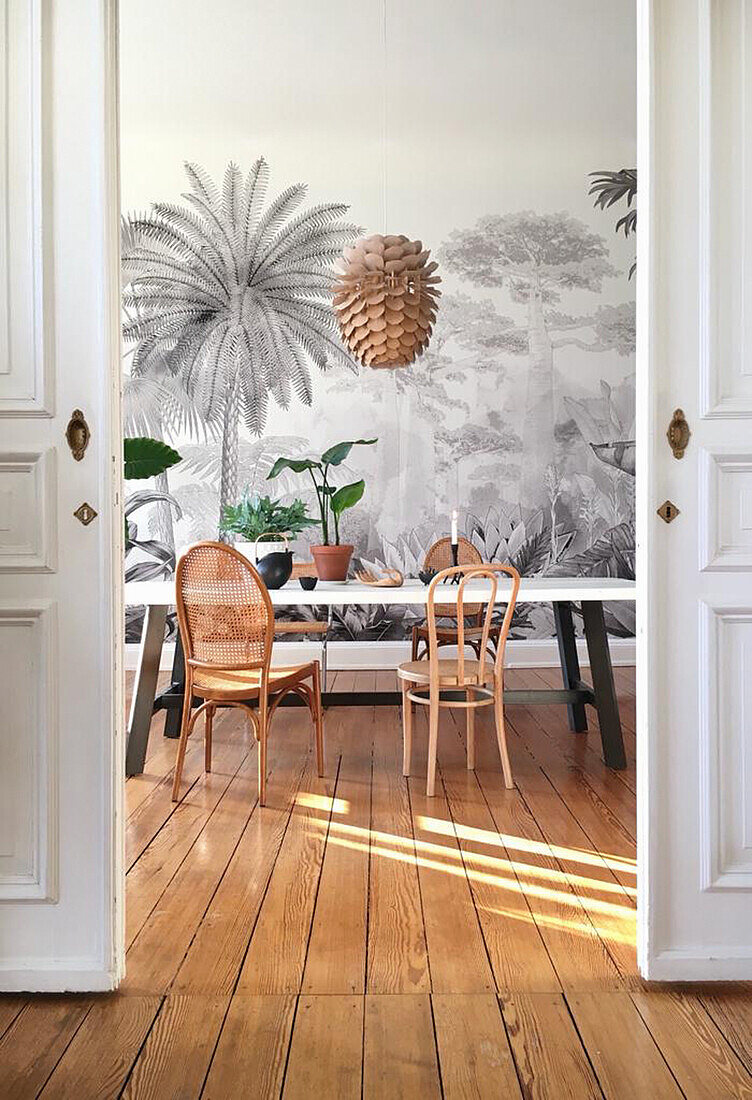 Table with various chairs and houseplants in dining room with mural wallpaper and wooden floorboards