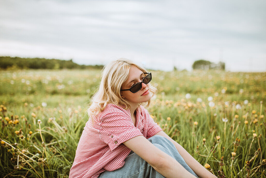 Side view of young serene female in trendy outfit sitting in blossoming meadow in summer and looking away