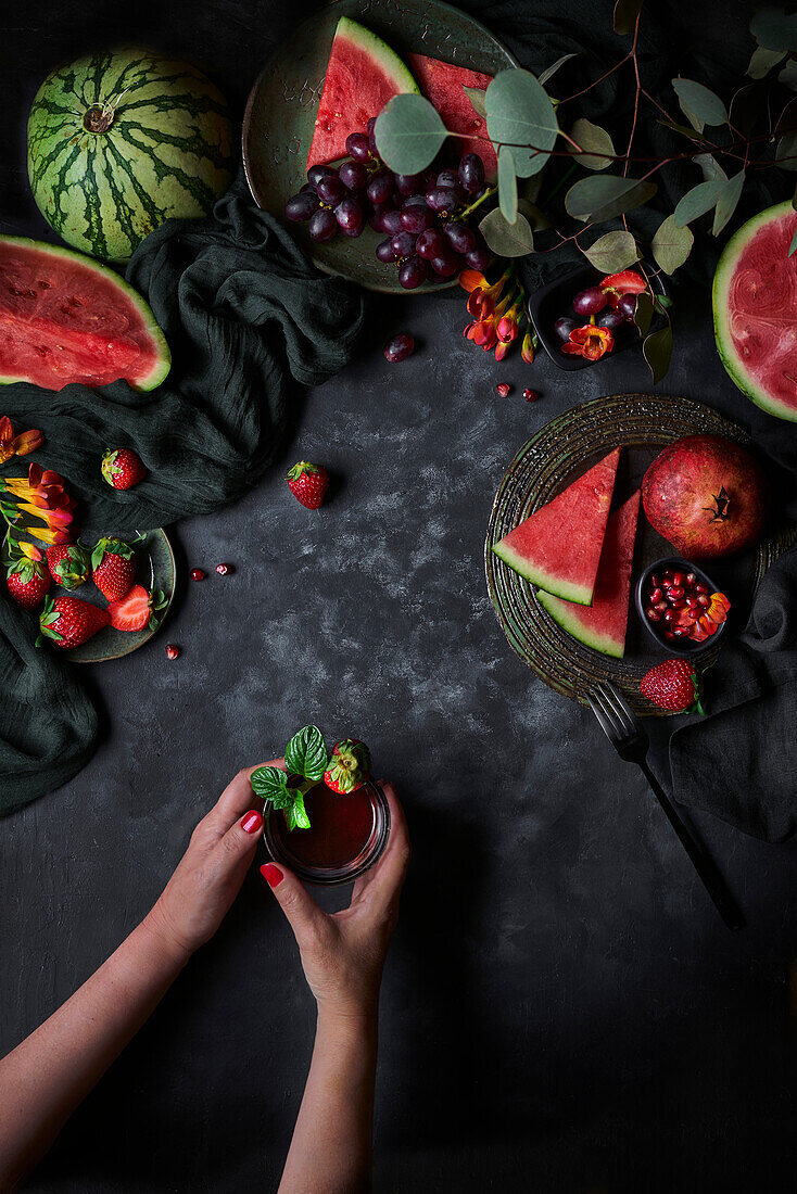 Glass of beverage topped with strawberry and basil leaves on black background with watermelon and pomegranate