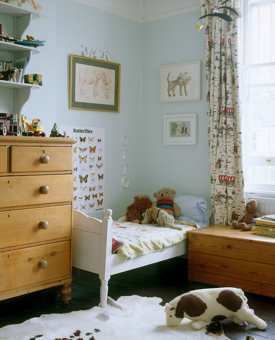 Ein traditionelles Kinderzimmer mit einer Reihe von Schubladen aus Kiefernholz neben einem kleinen Bett, umgeben von gerahmten Bildern und Postern und einer Holztruhe unter dem Fenster