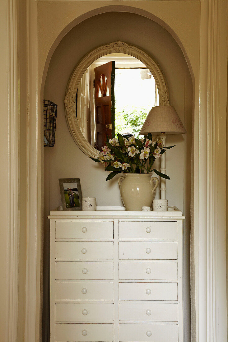 Cut flowers and oval shaped mirror on chest of drawers in recessed alcove of hallway in West Sussex home, England, UK