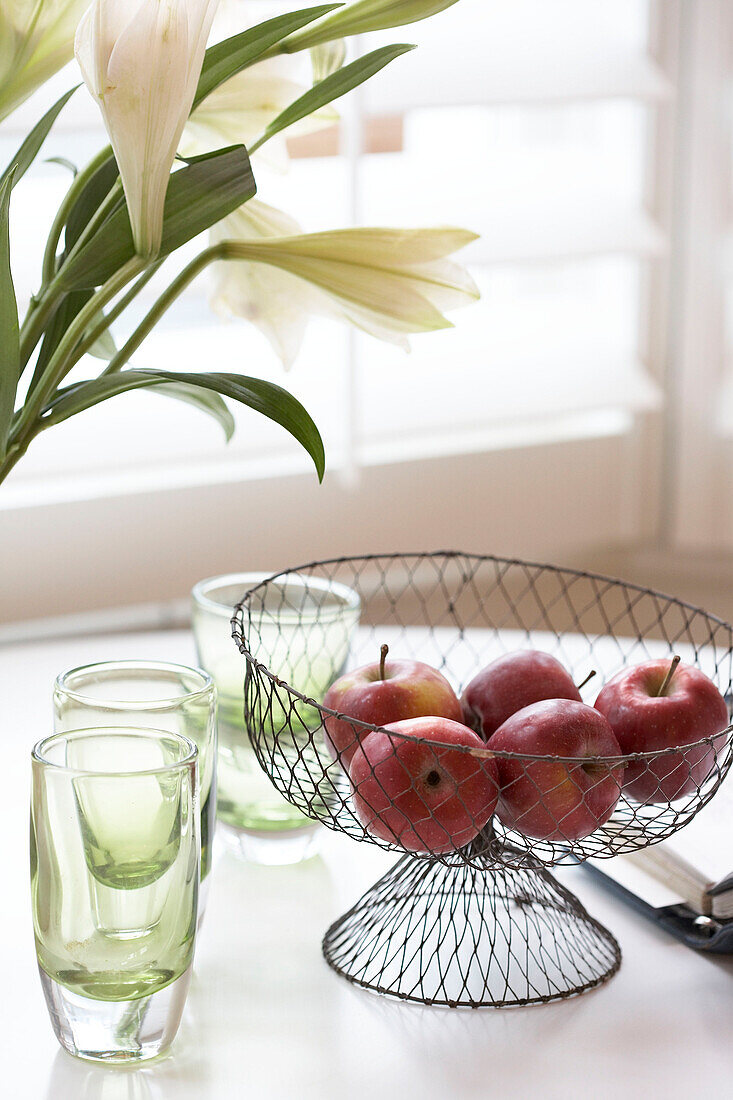 Close up detail of metal framed bowl of apples next to several coloured glasses overlooked by flowers