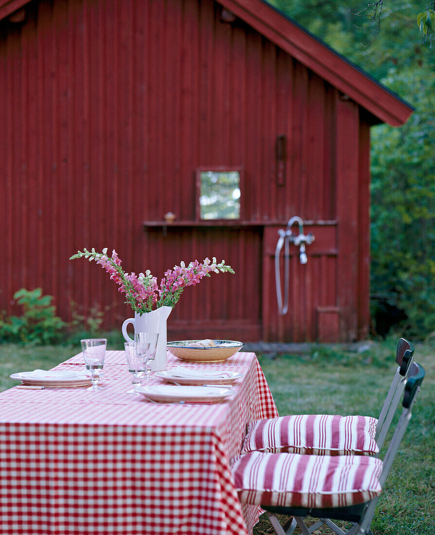 An outside detail of a table set for lunch in a garden chairs with pink striped cushions wooden house in the background