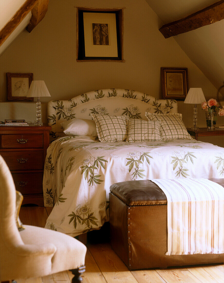 A traditional attic bedroom with beamed ceiling double bed with floral bedcover leather ottoman wooden flooring