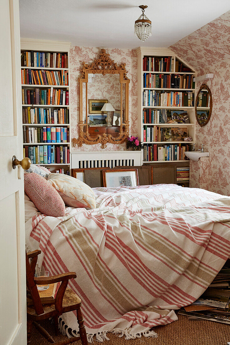 Unmade bed with bookshelves and Toile de Jouy wallpaper in 17th century Hampshire home, UK