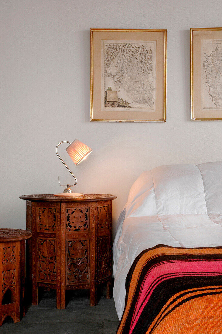 Lit lamp on carved bedside table with striped bed cover below cartographic artwork