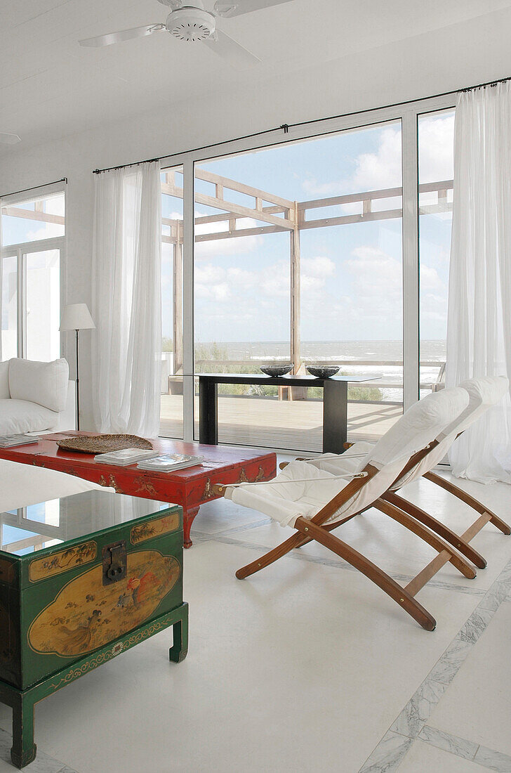 Huge windows overlooking the sea with linen curtains and a Chinese wood table lacquered in red with golden motifs sofas and deck-chairs in white canvas