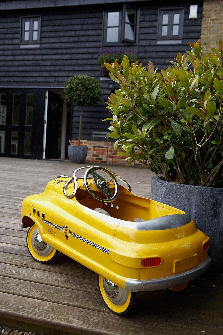 Bright yellow child's toy pedal car taxi on garden deck beside a pot plant