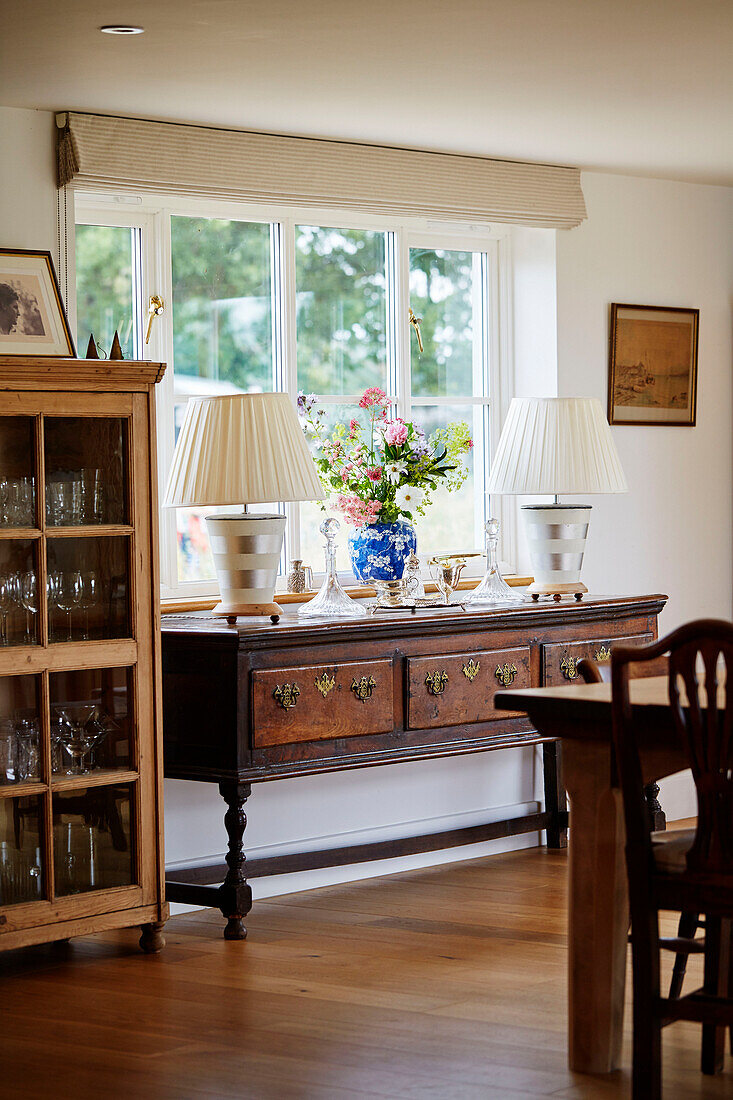Pair of lamps on wooden sideboard with glassware cabinet in Oxfordshire farmhouse, UK