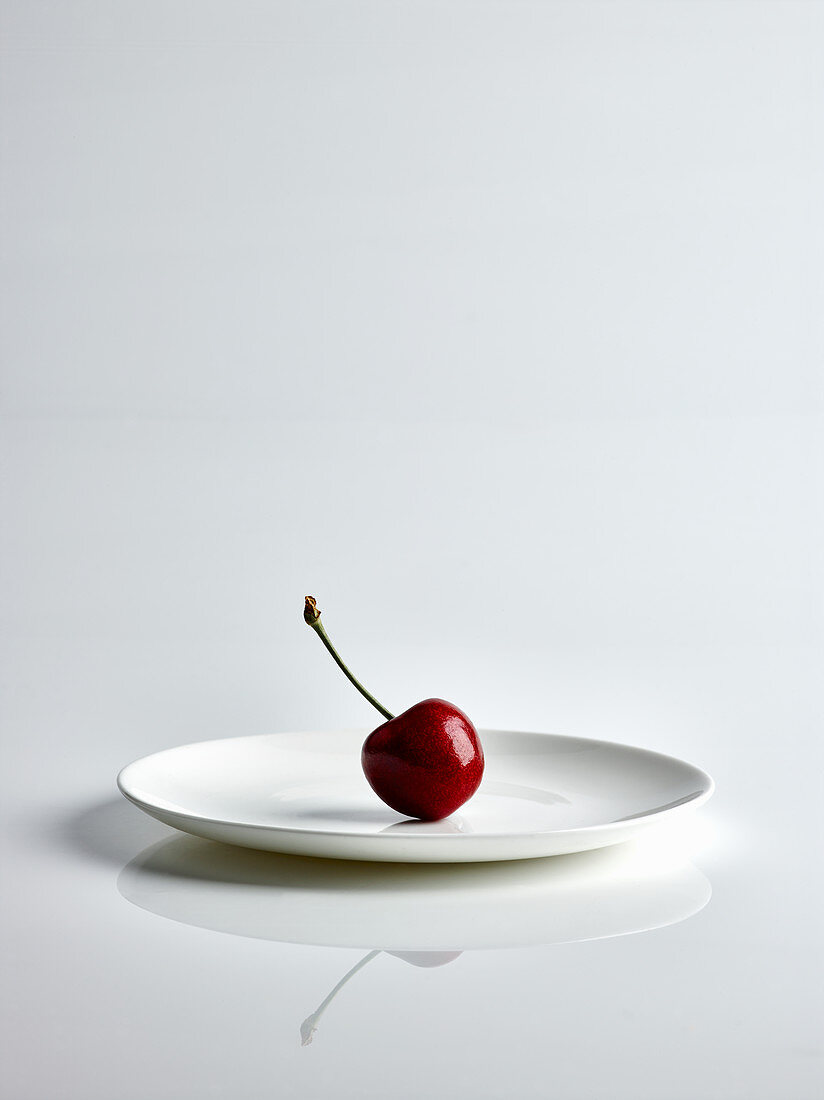 Eine Kirsche auf weißem Teller vor weißem Hintergrund