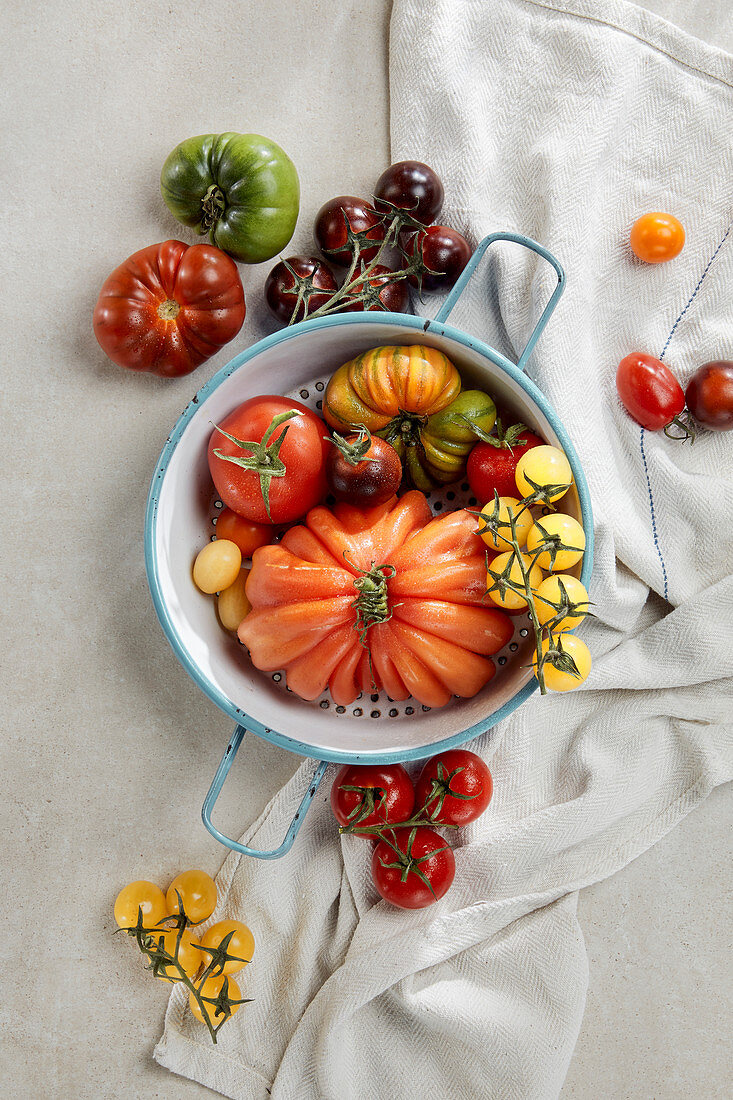 Various colourful tomatoes on a stone surface with an enamel colander and a tea towel