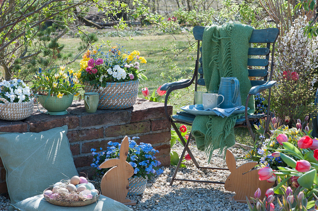 Small gravel terrace in the garden with Easter eggs and wooden Easter bunnies, chair with blanket, cups, jug and glasses